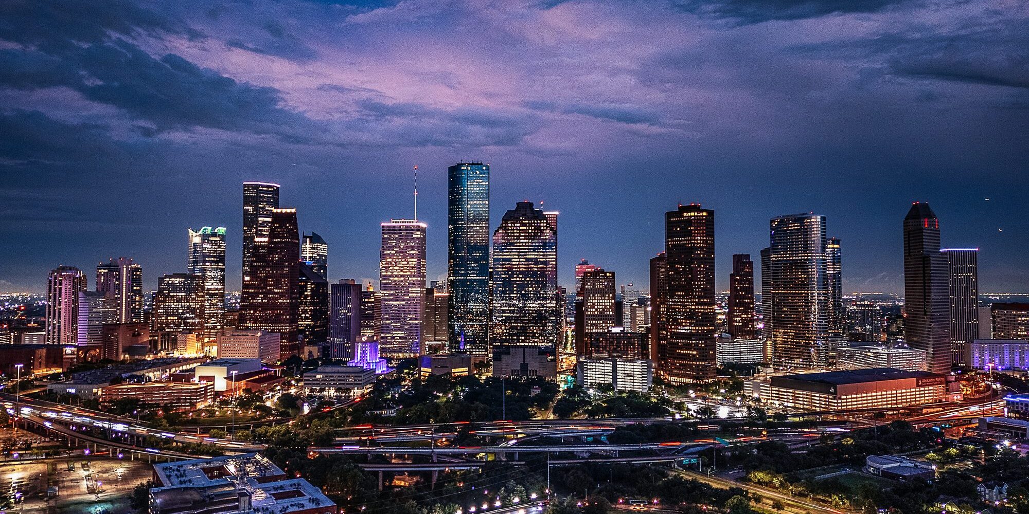 Drone aerial image of downtown Houston, Texas captured at twilight. The photograph highlights the illuminated skyscrapers, highways, and cityscape under a dramatic evening sky. A vibrant view of one of the largest cities in the United States and a major economic hub.